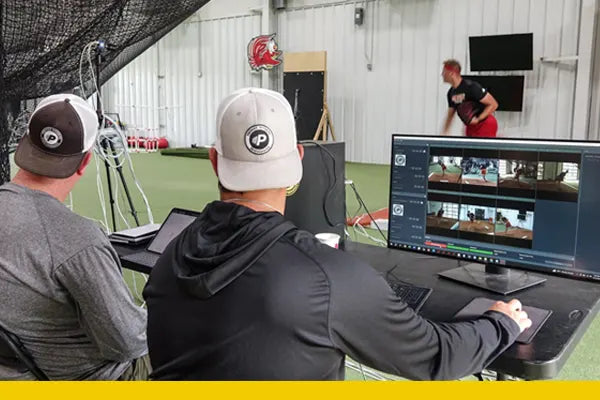 Two coaches analyze pitching footage and data on monitors during a comprehensive assessment session.