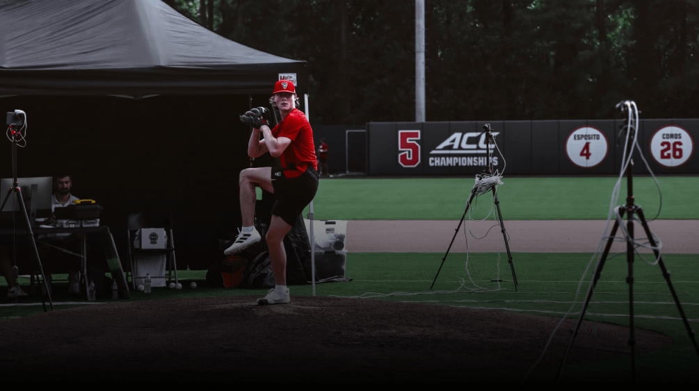 A baseball pitcher in a red uniform prepares to throw during a Premier Collegiate Development League session.